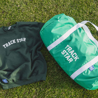 Green Track Star duffle bag on the ground next to the Track Star crewneck