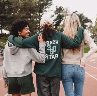 Three girls with arms around each other walking on a track with the girl in the middle wearing the Track Star hoodie